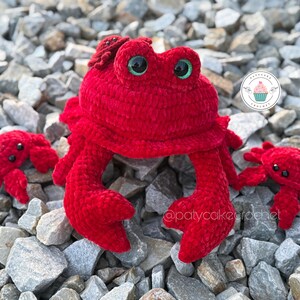 Pode incluir: Três caranguejos vermelhos de crochê, um grande e dois pequenos, em um leito de cascalho cinza. O caranguejo grande tem olhos verdes grandes e um pequeno caranguejo vermelho na cabeça.