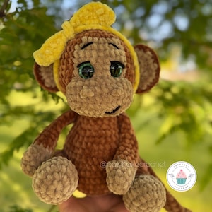 May include: A handmade crochet monkey doll in shades of brown and tan, with a yellow headband. The monkey has green eyes and is holding its legs in a sitting position. The doll is soft and plush, perfect for a child's toy or nursery decor.