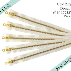 May include: Five gold zipper pulls with a donut shape. The zippers are 6, 8, 10, 12, and 14 inches long. The zippers are white with gold teeth and a gold pull.