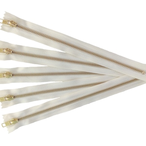 May include: Five white zipper tapes with gold metal teeth and sliders. The zippers are arranged in a fan shape.