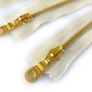 May include: Two white zipper tapes with gold metal zippers and pulls. The zipper pulls have a metal loop and the text "YKK" is visible on one of the pulls.