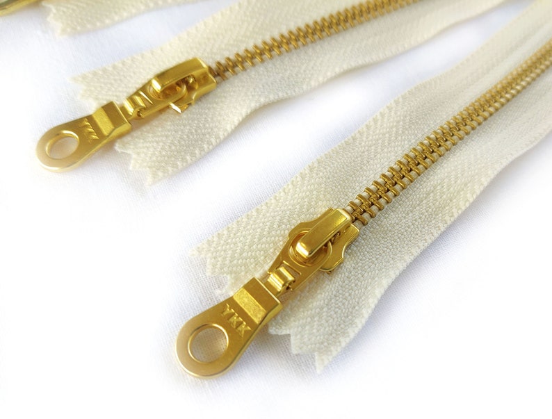 May include: Two white zipper tapes with gold metal zippers and pulls. The zipper pulls have a metal loop and the text "YKK" is visible on one of the pulls.