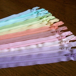 Puede incluir: Un surtido arco&iacute;ris de cinta de cremallera en varios colores pastel, incluyendo azul claro, verde, amarillo, naranja, rosa y morado. Las cremalleras est&aacute;n dispuestas en forma de abanico, con el color m&aacute;s claro en la parte superior y el color m&aacute;s oscuro en la parte inferior.