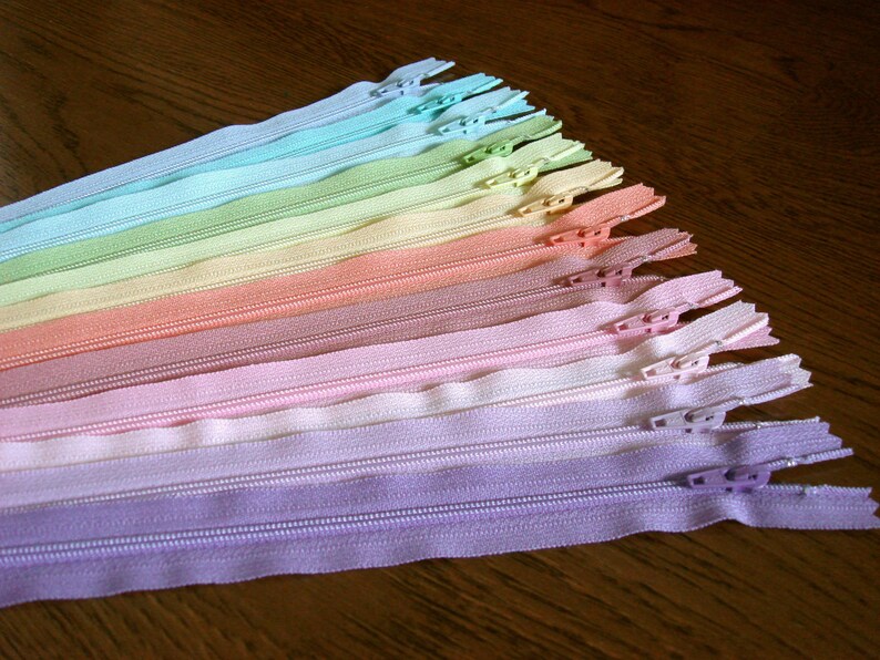 Puede incluir: Un surtido arco&iacute;ris de cinta de cremallera en varios colores pastel, incluyendo azul claro, verde, amarillo, naranja, rosa y morado. Las cremalleras est&aacute;n dispuestas en forma de abanico, con el color m&aacute;s claro en la parte superior y el color m&aacute;s oscuro en la parte inferior.