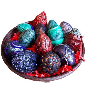 Puede incluir: Una colección de huevos de dragón en un cuenco marrón. Los huevos están hechos de cerámica y están pintados en varios colores, incluyendo rojo, azul, verde y morado. Los huevos están decorados con escamas y otros detalles.