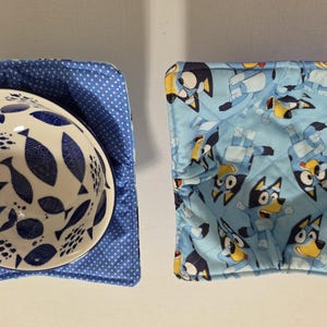 May include: Two bowl cozies. One is blue with white polka dots, holding a white bowl with a blue fish pattern. The other is light blue with a cartoon dog pattern. These cozies are designed to hold hot bowls.