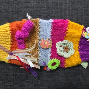May include: A colorful, handmade knitted toy with various textures and embellishments. The toy features stripes of yellow, orange, brown, pink, and purple. It includes ribbons, buttons, a green hair tie, and a wooden flower and cat-shaped button.