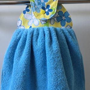 Op de afbeelding: Een blauwe handdoek van badstof met een gele en blauwe bloemenprint hanglus. De lus heeft een gele knop.