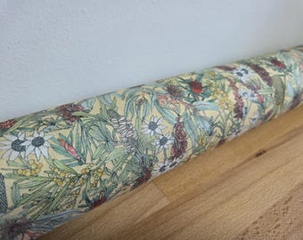 May include: Rolled fabric with a botanical print featuring various flowers and foliage in shades of green, red, white, and yellow. The fabric is on a light yellow background and rests on a wooden surface.