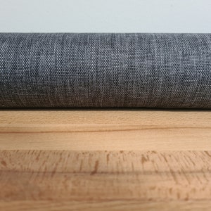 May include: Close-up of a gray fabric roll with a textured weave. The fabric is lying on a light brown wooden surface.