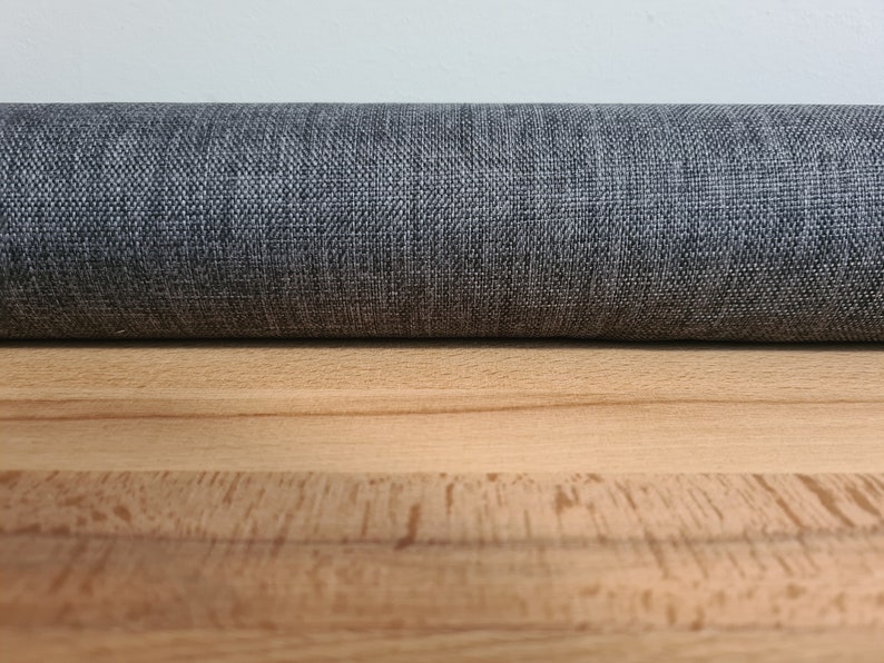 May include: Close-up of a gray fabric roll with a textured weave. The fabric is lying on a light brown wooden surface.