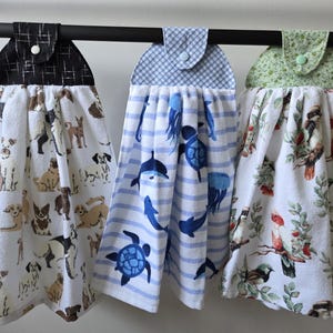 Puede incluir: Tres paños de cocina colgantes con la parte superior decorativa. Uno presenta un patrón de perros, otro tiene criaturas marinas azules sobre rayas y el tercero tiene un diseño de pájaros y flores. Cada paño está sujeto a una barra negra.