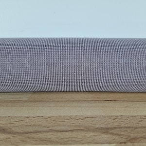 Può includere: Primo piano di un materiale a rete testurizzato di colore lilla chiaro. Il materiale è arrotolato e si trova su una superficie di legno marrone chiaro.