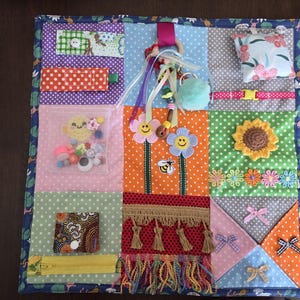 May include: A square sensory activity quilt featuring a grid of colourful fabric patches with various textures and attachments, including a clear pocket with beads, a wooden ring with ribbons, a sunflower applique, fringe, and small bows.