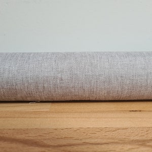 May include: A close-up of a light grey fabric with a subtle woven texture. The fabric is rolled up and resting on a wooden surface.