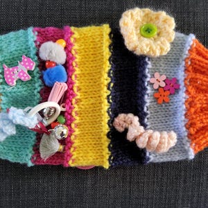 Puede incluir: Un juguete sensorial tejido a mano, colorido y adornado con diversas texturas y decoraciones. Incluye una flor tejida, pompones, campanas y un botón rosa en forma de perro. El juguete está diseñado con múltiples colores y texturas para la exploración táctil.