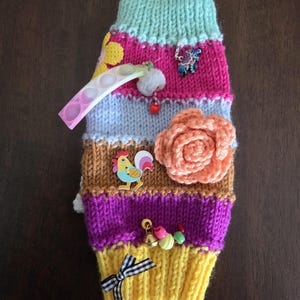 May include: A multi-colored knitted sweater for a small animal featuring horizontal stripes in mint green, pink, light blue, brown, purple, and yellow. It is decorated with a crochet flower, a rooster charm, a pop-it toy, and a small bow.