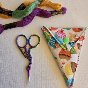 May include: An image of craft supplies, including green, yellow, and purple embroidery threads. A pair of iridescent embroidery scissors and a cupcake-patterned needle case are also visible. The items are arranged on a white surface.