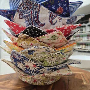 Puede incluir: Una pila de salvabols de tela hechos a mano en varios patrones y colores. Los salvabols presentan diseños florales, de cachemira y de estrellas. Los salvabols están apilados sobre una superficie de madera. Diseñados para microondas.