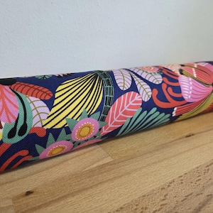 Puede incluir: Tela enrollada con un vibrante estampado floral. El diseño incluye grandes flores rosas, amarillas y rojas, hojas verdes y un fondo azul marino. La tela está sobre una superficie de madera.