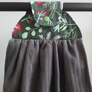 Op de afbeelding: Een grijze keukendoek met een patroon aan de bovenkant met rode bessen en groene bladeren op een donkergroene achtergrond. De handdoek hangt aan een zwarte stang, met een knoopsluiting.