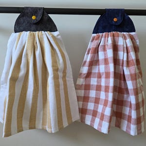 May include: Two hanging kitchen towels. One is white with yellow stripes, the other is orange and white checkered. Each towel has a dark gray top with a yellow button.