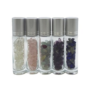 May include: Five clear glass roller bottles with silver lids filled with various coloured crystals. The bottles are lined up in a row from left to right. The crystals are white, pink, green, purple, and blue.