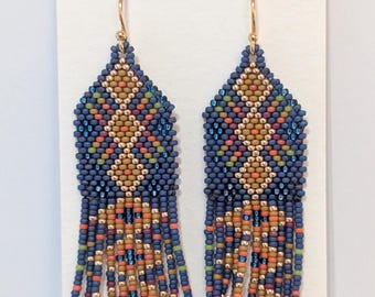 May include: A pair of beaded earrings with a geometric design. The earrings feature a dark blue background with diamond patterns in gold, orange, and other colors. The fringe is a mix of orange, blue, and green beads.