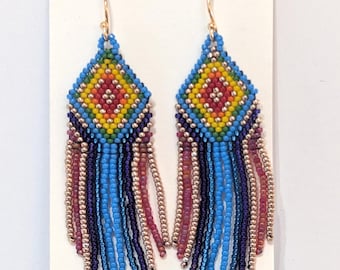 May include: Handmade beaded earrings with a diamond-shaped top in yellow, red, and blue, and long fringe in shades of blue, purple, and pink. The earrings hang from gold-colored hooks, showcasing a vibrant, colorful design.