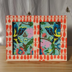 Peut inclure: Un cadre photo double rectangulaire avec un motif floral. Le cadre a une base crème avec un motif répétitif de gouttes orange. L'intérieur présente un fond turquoise avec un motif floral coloré en bleu, rose et jaune.