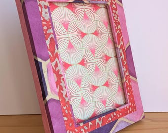 Puede incluir: Un marco de fotos rectangular con un diseño estampado en rosa, morado y rojo. El marco tiene una construcción en capas con una abertura central que muestra un patrón en forma de abanico en rosa y blanco. El marco está en ángulo, mostrando su superficie texturizada.