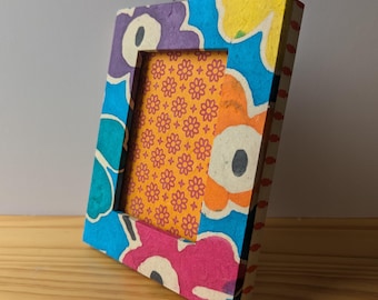 May include: A colorful picture frame made of recycled paper with a floral design. The frame is blue, yellow, orange, pink, and purple. The frame is empty and ready for a photo.