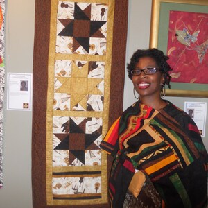K&ouml;nnte beinhalten: Eine Frau in einem bunten Patchwork-Schal steht vor einer Wand mit zwei gerahmten Kunstwerken. Das Kunstwerk links ist eine Steppdecke mit braunem Hintergrund und einem sich wiederholenden Muster aus Sternen und Musikinstrumenten. Das Kunstwerk rechts ist ein abstraktes rotes und goldenes Gem&auml;lde mit einem blauen und gr&uuml;nen Vogel.