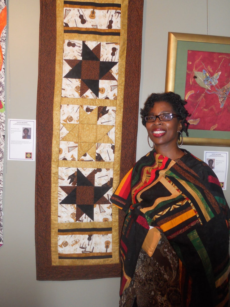 K&ouml;nnte beinhalten: Eine Frau in einem bunten Patchwork-Schal steht vor einer Wand mit zwei gerahmten Kunstwerken. Das Kunstwerk links ist eine Steppdecke mit braunem Hintergrund und einem sich wiederholenden Muster aus Sternen und Musikinstrumenten. Das Kunstwerk rechts ist ein abstraktes rotes und goldenes Gem&auml;lde mit einem blauen und gr&uuml;nen Vogel.