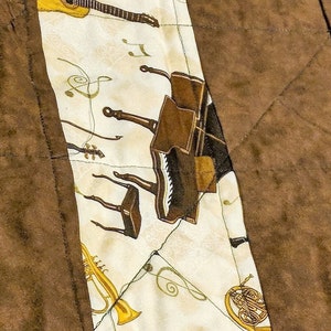 K&ouml;nnte beinhalten: Ein wei&szlig;er Stoff mit aufgedruckten Musikinstrumenten. Die Instrumente umfassen eine Gitarre, eine Harfe, ein Klavier, eine Trompete und ein Waldhorn. Der Stoff ist an einen braunen Stoff gen&auml;ht.