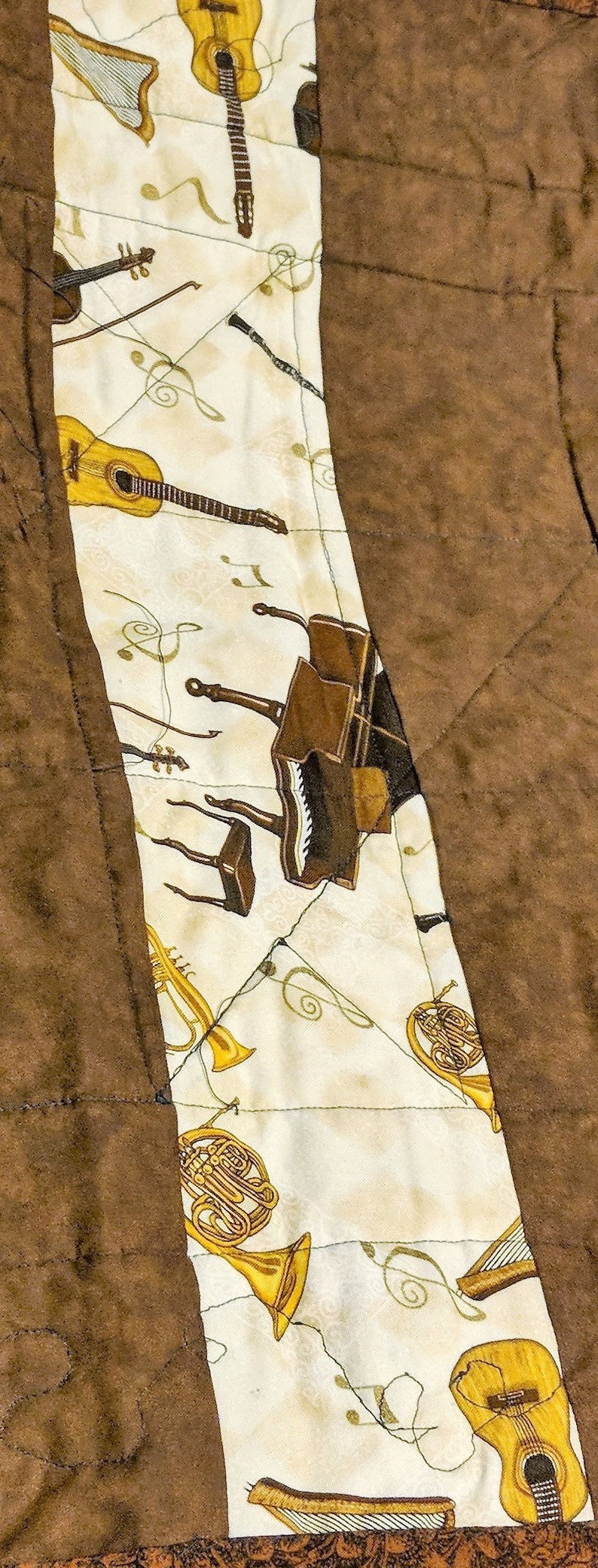K&ouml;nnte beinhalten: Ein wei&szlig;er Stoff mit aufgedruckten Musikinstrumenten. Die Instrumente umfassen eine Gitarre, eine Harfe, ein Klavier, eine Trompete und ein Waldhorn. Der Stoff ist an einen braunen Stoff gen&auml;ht.