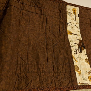 K&ouml;nnte beinhalten: Eine braune gesteppte Wurfdecke mit einem wei&szlig;en Stoffpanel, das Musikinstrumente zeigt, darunter eine Gitarre, ein Klavier, eine Trompete und eine Fl&ouml;te.