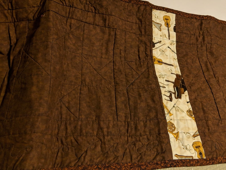 K&ouml;nnte beinhalten: Eine braune gesteppte Wurfdecke mit einem wei&szlig;en Stoffpanel, das Musikinstrumente zeigt, darunter eine Gitarre, ein Klavier, eine Trompete und eine Fl&ouml;te.