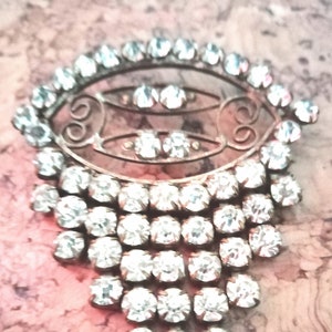 May include: A silver brooch with a delicate filigree design and a cluster of clear rhinestones. The brooch features a teardrop shape with a cascading row of rhinestones.