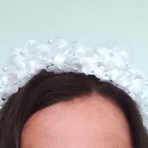 May include: A white floral headband with small pearl accents. The headpiece features delicate fabric flowers and small, round pearls. The headband is set against a light blue background, creating a soft, elegant look. This accessory is suitable for weddings or special occasions.