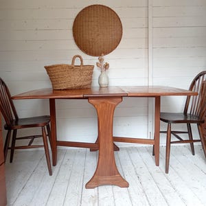 May include: A brown wooden drop-leaf table with a central pedestal base, flanked by two dark brown wooden spindle-back chairs. A woven straw basket and a small ceramic vase with dried stems sit on the table surface, with a round woven wall hanging.