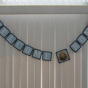 May include: A blue and white banner with the words "WELCOME HOME" hanging on a white slatted blind. A gold and blue emblem is in the center of the banner.