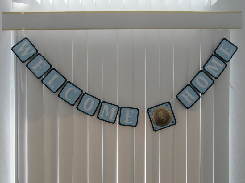 May include: A blue and white banner with the words "WELCOME HOME" hanging on a white slatted blind. A gold and blue emblem is in the center of the banner.