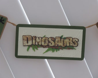 May include: A green and brown banner with the word "Dinosaurs" in brown letters with a camouflage pattern. The banner is hanging from a string.