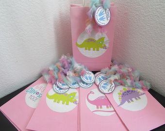 May include: Pink party favor bags with dinosaur designs and the text "Girls Dino Party Too!"  The bags are decorated with colorful dinosaur stickers and a pink and white feather boa.