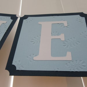 May include: A blue banner with a white letter "E" on it. The banner has a navy blue border and a white snowflake pattern.