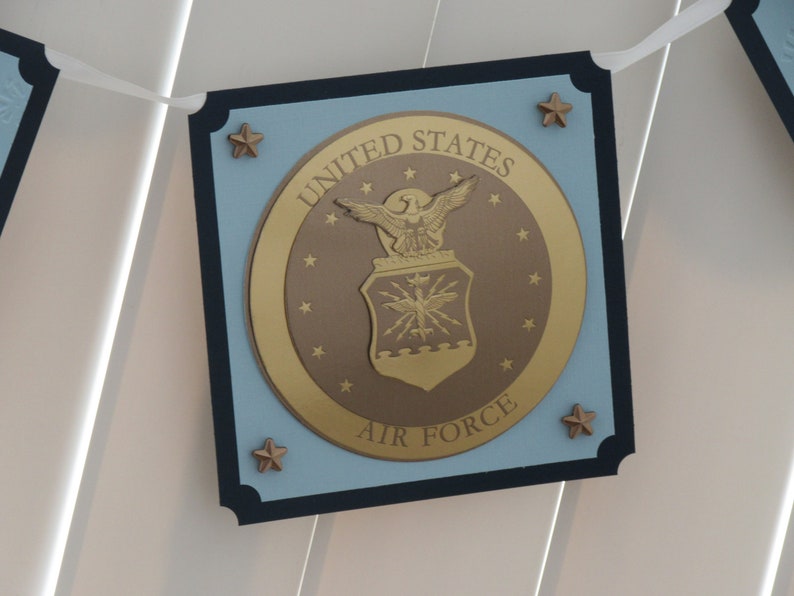 May include: A blue and black banner with a gold seal featuring the United States Air Force emblem. The seal has a bald eagle with outstretched wings and a shield with a star and lightning bolts. The text "UNITED STATES AIR FORCE" is written around the seal.