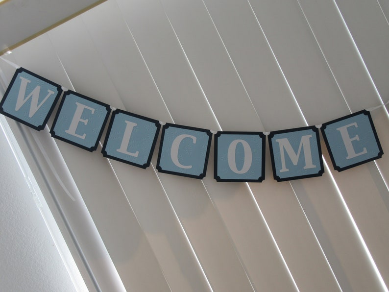 May include: A blue and black welcome banner with white letters. The banner is made of individual squares with a black border and a light blue background. The letters are white and are in a simple font.