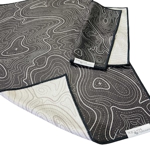 May include: A set of three dark gray microfiber towels with a white topographical map design. The towels have a black border and the text "PROJECT HOME" on a white label. The towels are folded and arranged on a white surface.