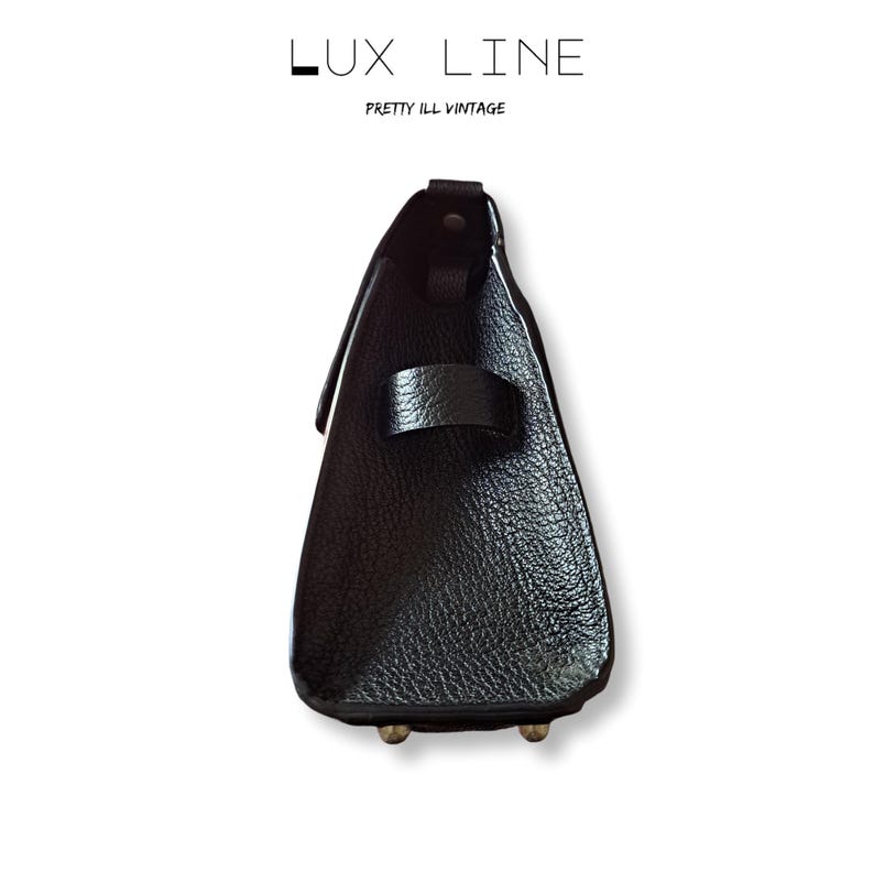 Puede incluir: Un bolso de mano de cuero negro con una superficie texturizada. El bolso tiene una correa y dos peque&ntilde;os pies met&aacute;licos. El texto "LUX LINE" y "PRETTY ILL VINTAGE" est&aacute; impreso en la parte superior.